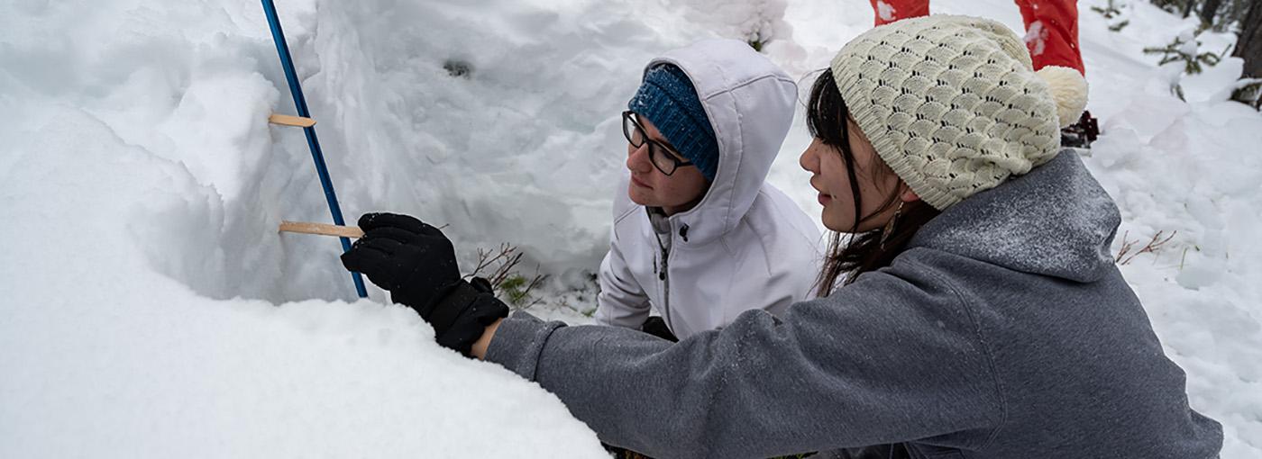 Two students use popsicle sticks to measure the distance between snow layers in a snow pit.